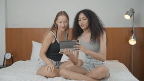 Two diverse woman sitting on bed watching video or clip video on digital tablet.. Young girl spending time with girlfriend on weekend at home