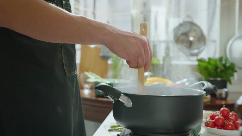 Male chef in apron stirring boiling food in a pot with wooden spatula while preparing dinner in the kitchen. Cropped shot, side view Royalty-Free Stock Footage #1108855167