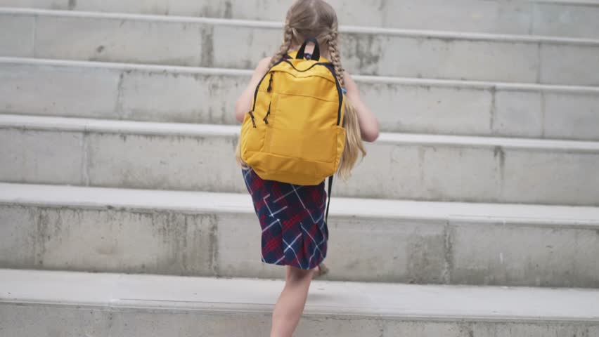 Little girl with yellow schoolbag goes up stone steps in modern city schoolgirl with backpack on high stairs backside view child student goes to school