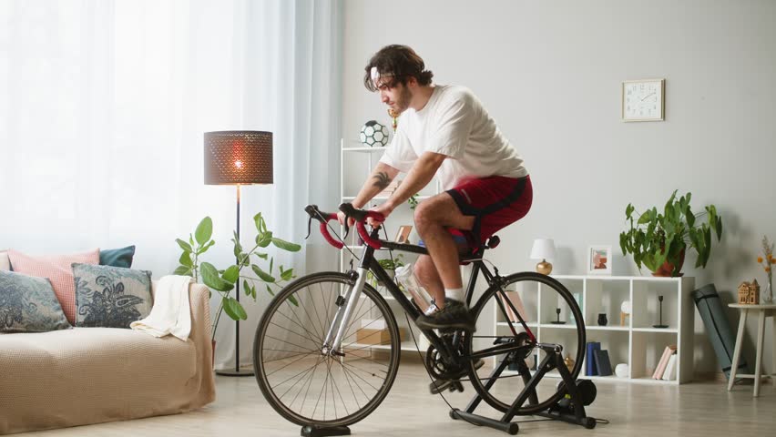 Sportsman riding bicycle, home gym. Young man cycling in living room, exercising in morning. Spanish guy preparing for competition, work out on stationary bike. Healthy lifestyle.  | Shutterstock HD Video #1108858321