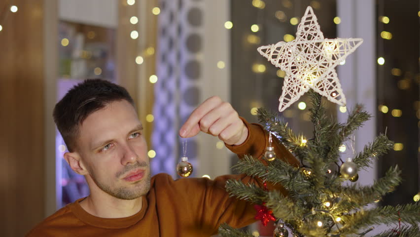 Upset middle-aged man sadly twirls small glass decorative ball and sighs. Lonely guy sits near Christmas tree and is disappointed with his situation. Lack of positive mood during holiday. | Shutterstock HD Video #1108859453