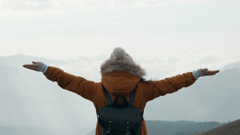 Стоковое видео: Happy asian woman tourist raise hands enjoys breathing fresh air high in cloudy mountains at cold weather. Young hiker at amazing hazy background of mountain landscape standing back. Active sport