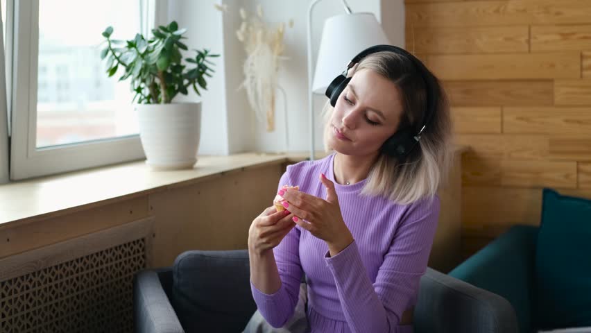 A beautiful blonde spends time alone at home. The girl sits in a chair by the window, listens to music on headphones and eats sweet cakes.