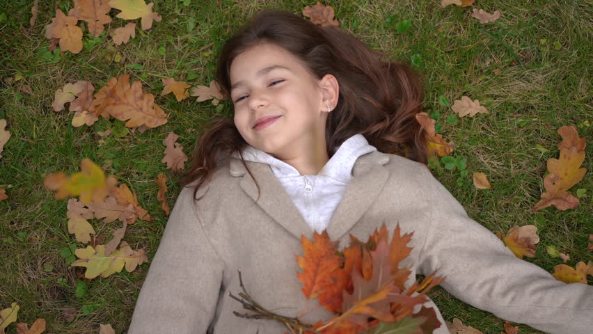 Top view carefree smiling girl lying on meadow as autumn leaves falling in slow motion. Portrait of relaxed Caucasian child enjoying tranquility in park outdoors on lawn