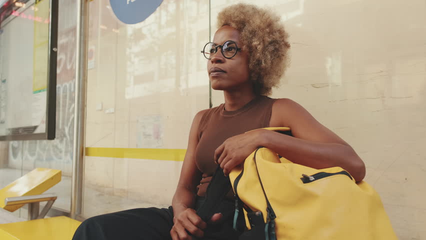 Young woman in glasses, dressed in brown top with backpack, sits at public transport stop looks around