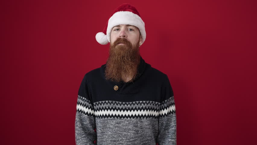 Young redhead man holding gift wearing christmas hat over isolated red background
