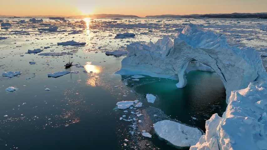 Small yacht sails between icebergs in the rays of sunset. Ship passes near huge ice cave in iceberg in Ilulissat, Greenland. Traveling on a yacht in the Arctic Circle, aerial shot Royalty-Free Stock Footage #1108874921