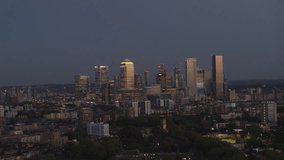 Drone video of Canary Wharf, shot from Bethnal Green, facing south east with drone moving forwards. City skyscrapers lit up with office lights and an orange sunset reflecting off the glass.