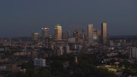 Drone video of Canary Wharf, shot from Bethnal Green, facing south east with drone moving right. City skyscrapers lit up with office lights. Football pitch with players in foreground.