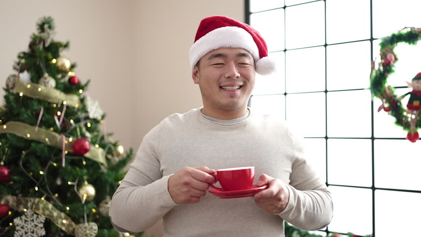 Young chinese man drinking coffee standing by christmas tree at home