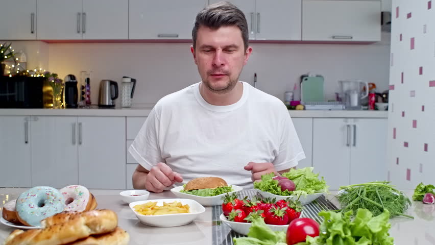 Man having unhealthy lunch at home in the kitchen, happy to eat fast food. Bulimia concept, full table of food and man eating alone