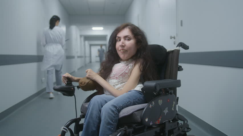 Woman with physical disability in electric wheelchair in the middle of clinic corridor smiles and looks at camera. Medical personnel walk in hallway of hospital or medical facility in the background. Royalty-Free Stock Footage #1108880687