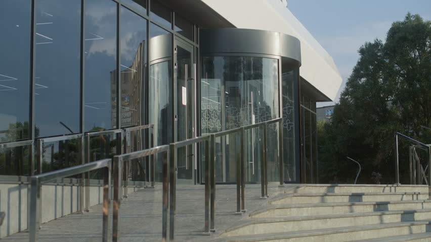Woman with SMA disability rides into clinic entrance on electric wheelchair. Medical worker meets female patient, helps her enter through revolving doors. Hospital or modern medical facility outside.