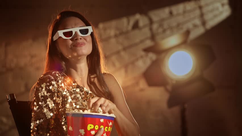 Closeup shot of attractive woman in golden dress wearing 3d paper glasses, watching comedy laughing and eating popcorn from bucket.