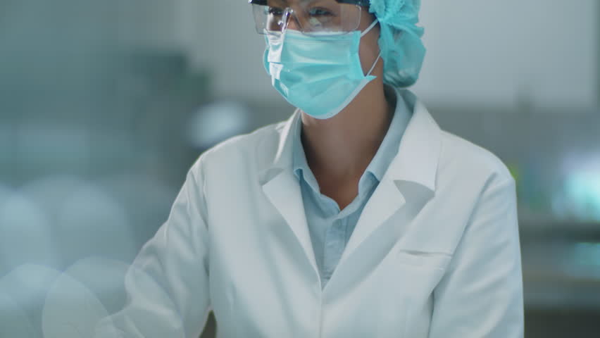 Asian female chemist in protective face mask, medical hat and gloves examining vial with chemical during laboratory research | Shutterstock HD Video #1108886669