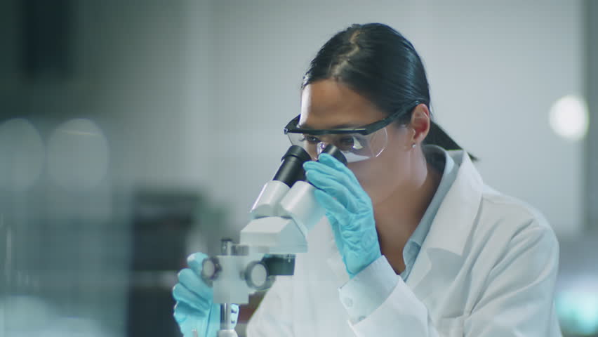 Medium shot of Asian woman scientist in medical gloves, protective eyewear and white coat looking into microscope and taking notes during laboratory experiment | Shutterstock HD Video #1108886675