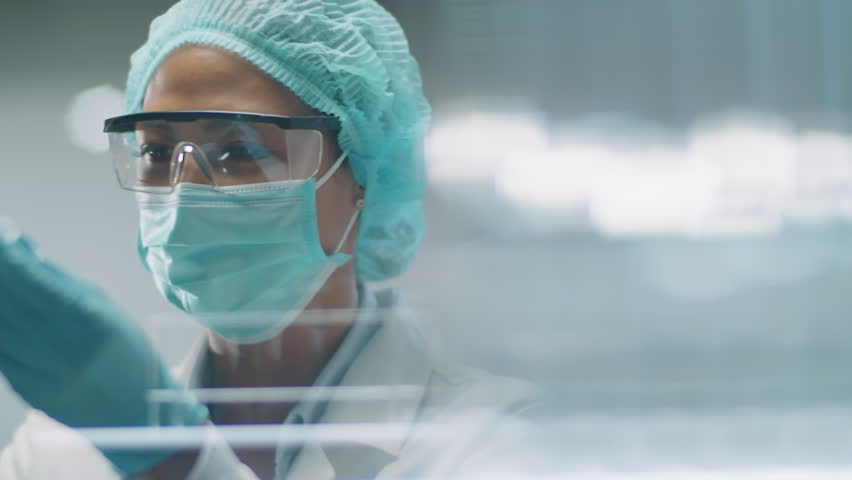 View through glass wall of Asian female scientist in protective mask, hat and gloves pouring blue chemical with pipette into test tube and looking at reaction during experiment in laboratory | Shutterstock HD Video #1108886695