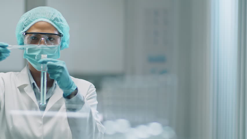 Medium shot of Asian female scientist in protective face mask, medical hat and gloves pouring blue chemical with pipette into test tube during laboratory experiment | Shutterstock HD Video #1108886701