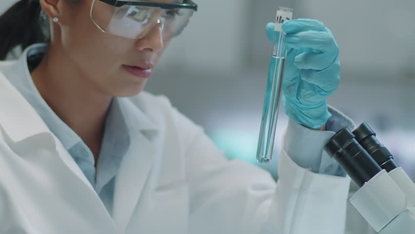 Asian female chemist in protective eyewear and medical gloves holding test tube and looking at blue liquid substance in it while working in laboratory | Shutterstock HD Video #1108886721