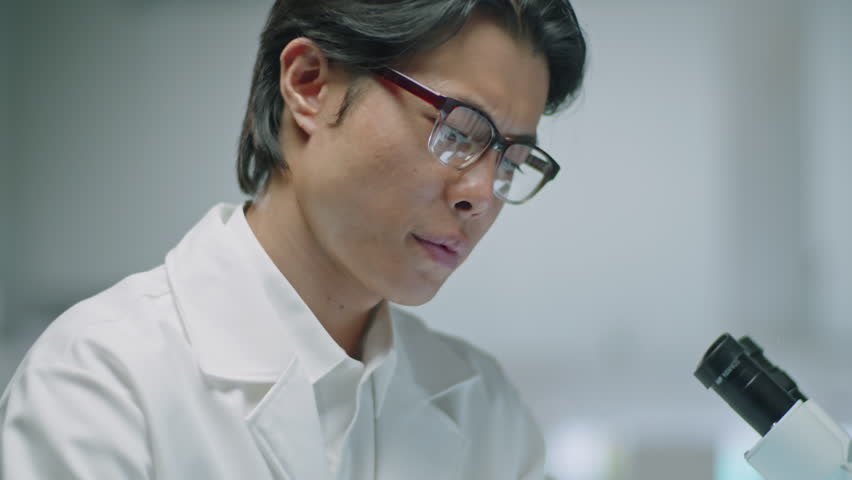 Tilt down shot of Asian scientist in lab coat and gloves writing down notes and taking test tube while working at desk in laboratory | Shutterstock HD Video #1108886749