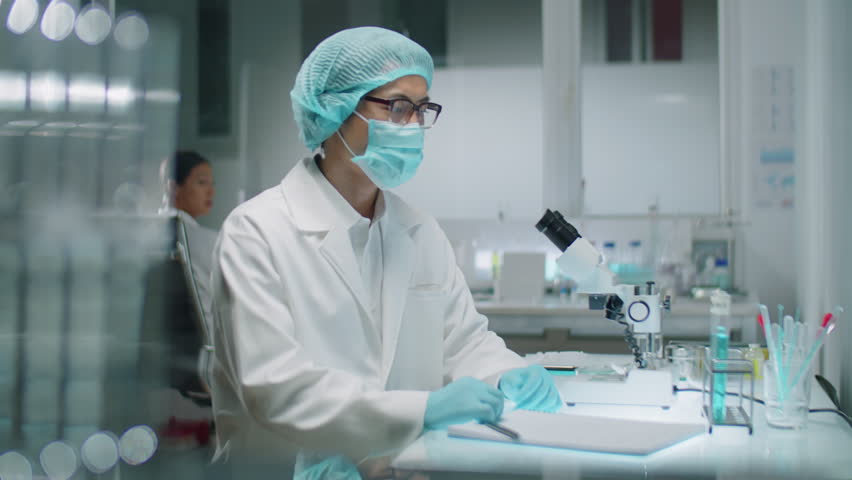 Medium shot of male chemist wearing protective face mask, medical hat and gloves examining liquid substance in test tube and writing notes while working in laboratory | Shutterstock HD Video #1108886761