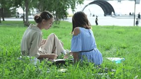 Cute happy little school children with backpacks and notebooks sitting in a grassy park outdoors. Two young schoolgirls doing homework outdoors
