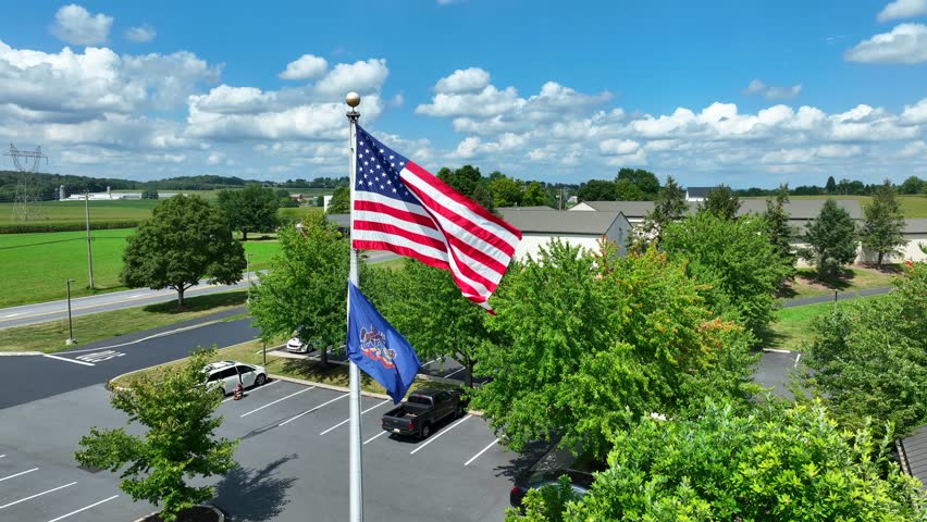 Pennsylvania and American flag waving in rural PA at local government building. Aerial rising shot revealing rolling hills and farmland on bright summer day.
