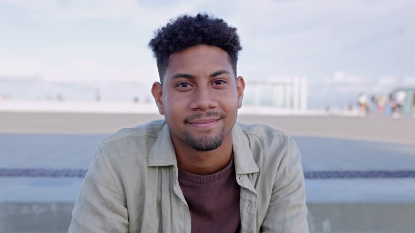 Young latin man smile at camera sitting outdoors. Smiling portrait of cheerful South American guy relaxing at city street. Slow motion footage, positive expression. Dominican people. Royalty-Free Stock Footage #1108892801