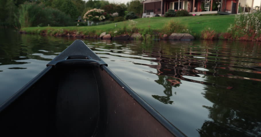 POV view of the boat nose floating along the river or lake and approaching the bank with nice green lawn and garden in front of the cosy house. Wonderful summer evening