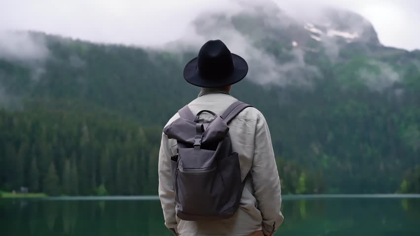 A young man with backpack with in a black hat looks at a forest lake. Advertising concept of hiking, advertising of clothes and accessories for tourism and travel. Amazing view high mountain lake 