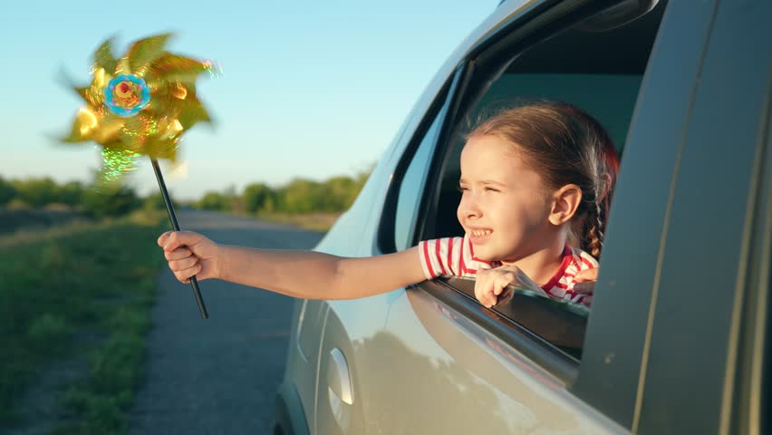 Cute little girl holds rotating windmill out of car window driving across farmland closeup. Little girl plays with windmill toy riding car with mother. Girl has fun with windmill during road trip Royalty-Free Stock Footage #1108899027