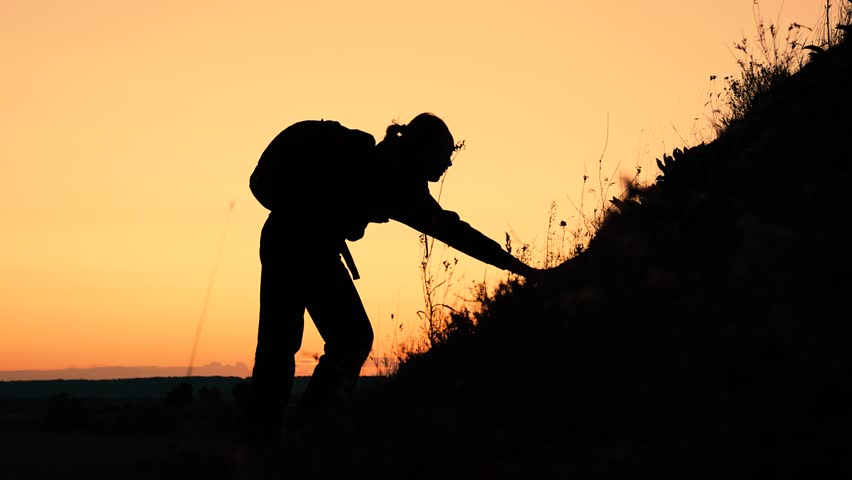Silhouette of woman with backpack climbing on hill with friend help at sunset. Woman climber gets support from friend on mountain slope. Woman tourist and friend teamwork during difficult climbing