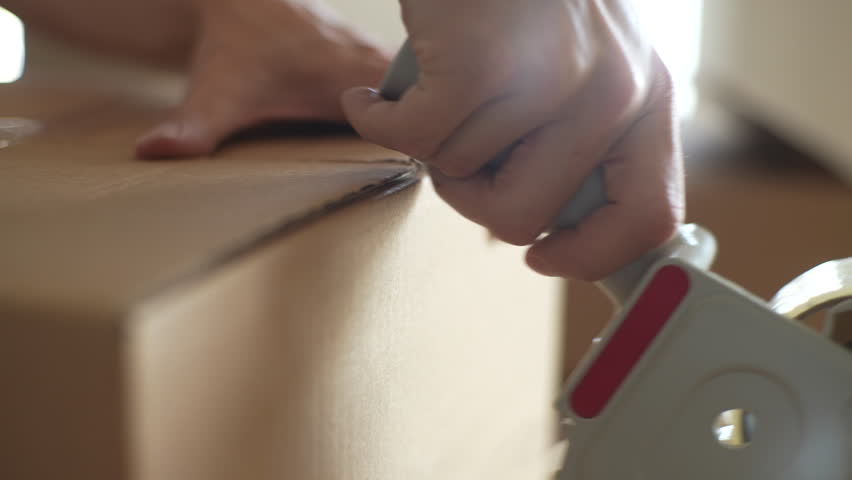 Close-up cropped shot of unrecognizable man packing up furniture and home belongings in boxes with adhesive roller for relocation. Packing boxes with adhesive tape for gluing cardboard boxes of goods,
