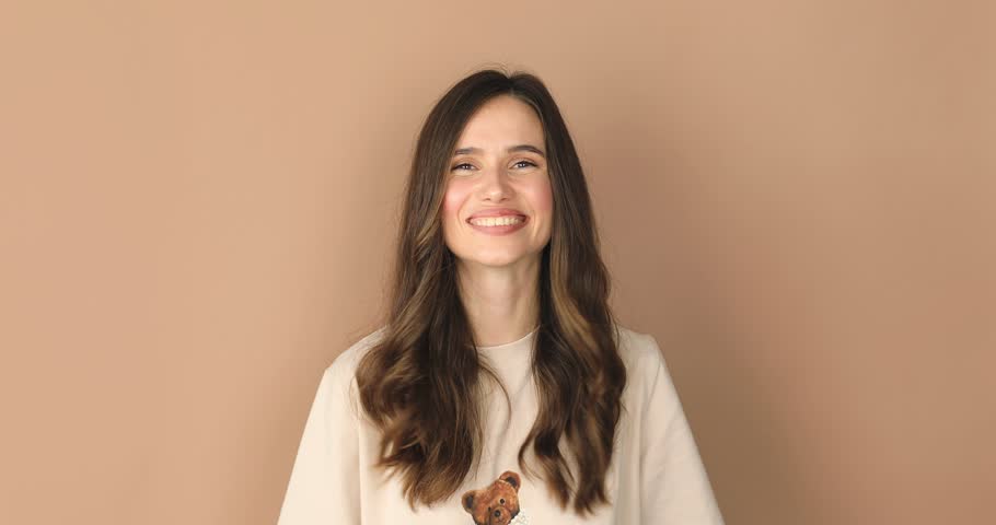 Young positive woman showing peace gesture, expressing fun and happiness, sign of victory, beige studio background. Young caucasian woman showing victory sign and smiling broadly.