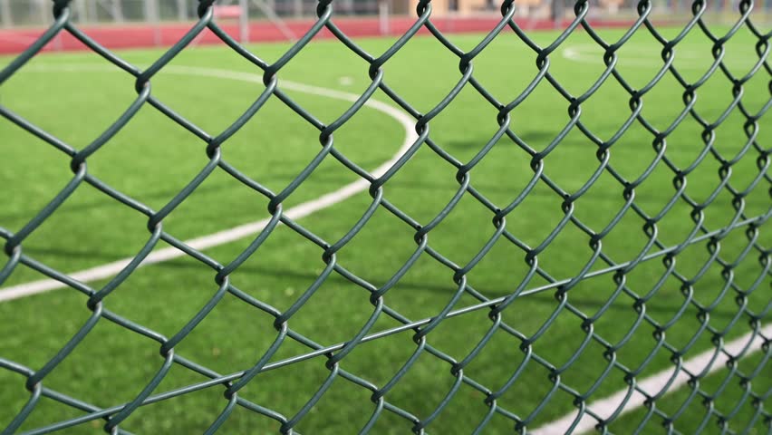 Through the chain-link fence, a blurred soccer field emerges, where passion for the game and dreams of victory come to life. Royalty-Free Stock Footage #1108903263