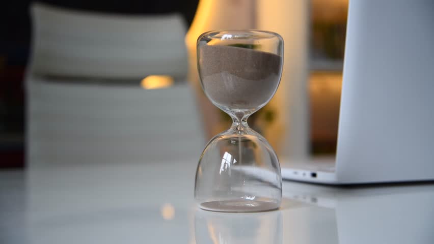 Close-up view of hourglass (sandglass or sand clock) standing on white table next to open laptop. Empty seat. Soft focus. Copy space. Real time video. Deadline theme. | Shutterstock HD Video #1108908081