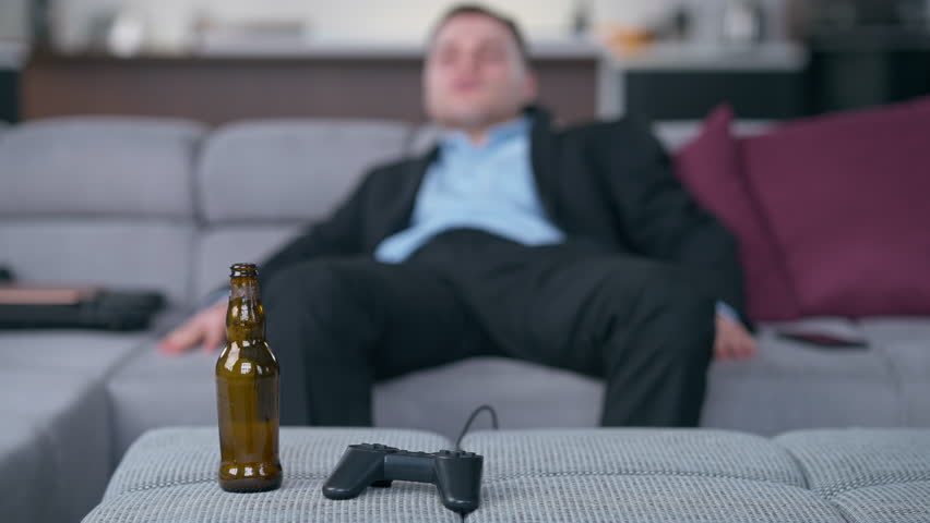 Empty beer bottle and joystick with blurred young drunk man lying on sofa at background. Caucasian unrecognizable depressed stressed businessman at home indoors | Shutterstock HD Video #1108911219