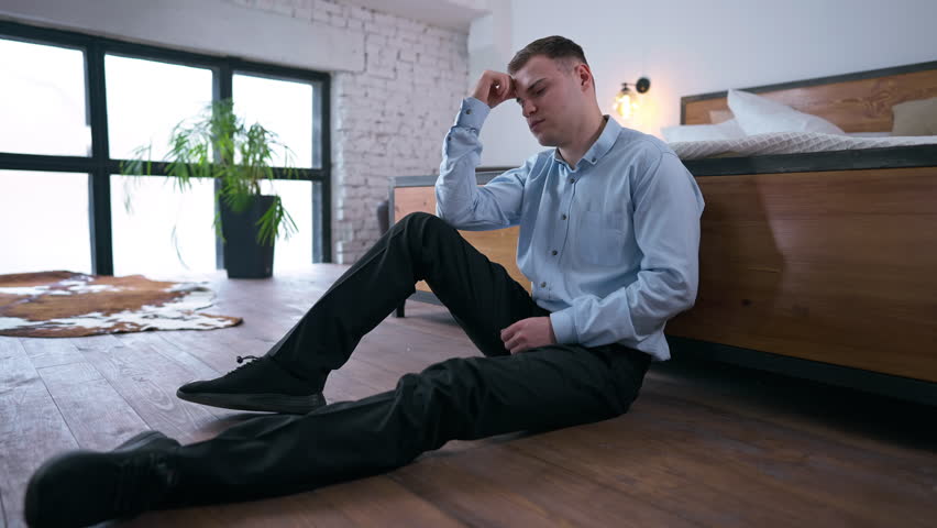 Wide shot stressed overburdened young businessman sitting on floor leaning on bed thinking. Side angle view portrait of tired worried Caucasian man looking away planning business startup | Shutterstock HD Video #1108911267