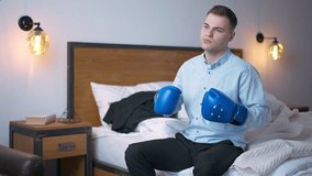 Medium shot portrait of confident young businessman hitting boxing gloves on hands in slow motion. Caucasian man looking away sitting on bed indoors thinking - Powered by Shutterstock - Get 15% off with code: PIKWIZARD15