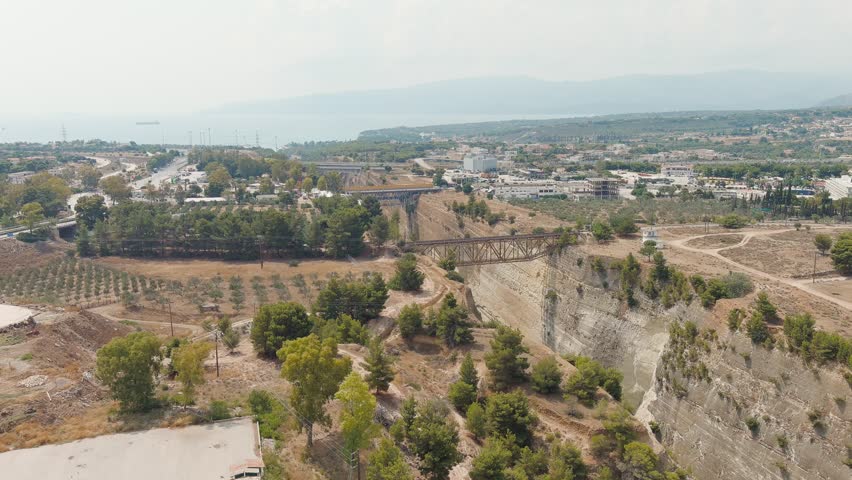 Corinth Canal, Greece. A narrow shipping canal cut through solid limestone rock connects the Saronic Gulf of the Aegean Sea with the Gulf of Corinth of the Ionian Sea. The waterway is flanked by steep, near-vertical rock walls rising approximately 90 metres above the waterline, creating a dramatic trench that separates the Peloponnese peninsula from the Greek mainland. The canal operates without locks, maintaining a constant sea level throughout its length. Several bridges span the cutting at height, including a prominent metal truss railway bridge and a modern road bridge visible in the distance. The surrounding landscape features dry, brown earth with scattered evergreen trees and low vegetation, alongside agricultural plots and a coastal town stretching toward the horizon. The water below glows turquoise against the pale limestone cliffs. Shot from an aerial perspective, the camera glides smoothly along the axis of the canal, revealing the full depth of the cutting and the scale of the engineering work. Bright daylight under a hazy sky gives the scene a warm, arid Mediterranean atmosphere typical of the Greek summer.