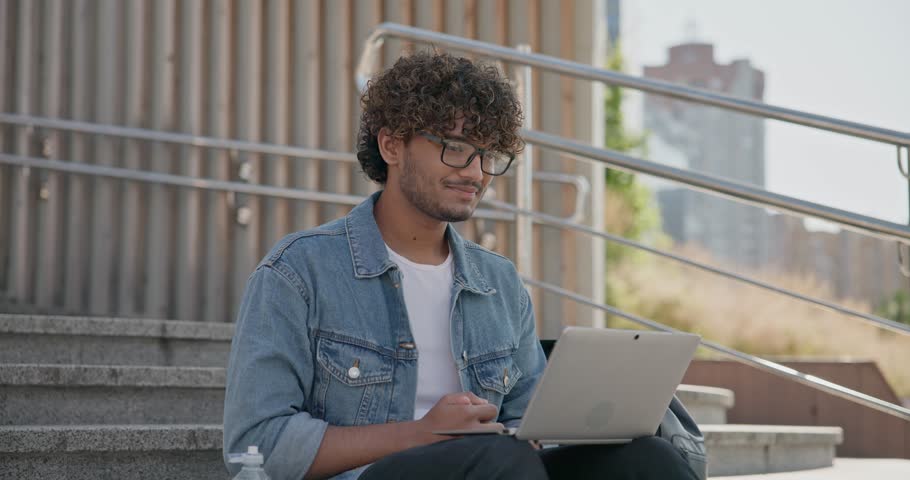 young handsome indian hindu man student freelancer sitting outdoors on steps making video call using laptop computer