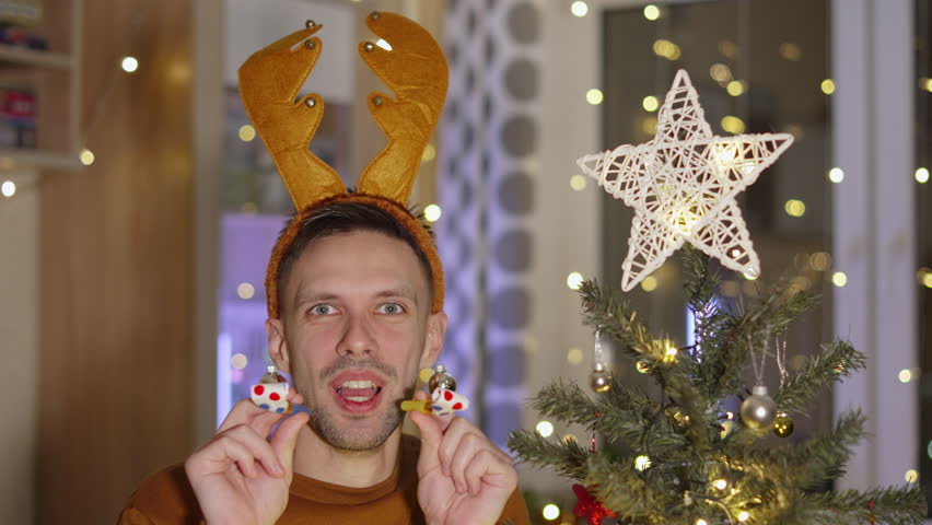 Funny brunette with toy deer antlers on head and glass balls of earrings blows two party horns at same time near Christmas tree in living room. Man loudly announces great news on eve of holiday. | Shutterstock HD Video #1108915809