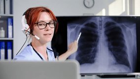 online medicine, cute female doctor in glasses talks on a video call using headset with patients using a web camera on laptop and shows the results of an X-ray while sitting in medical office