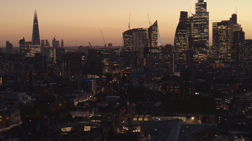 Aerial drone film near Bethnal Green, with the Shard, Gherkin and city lit internally and silhouetted against the orange sunset sky. Blue flashing lights from a police van can be seen on the street. Royalty-Free Stock Footage #1108926785