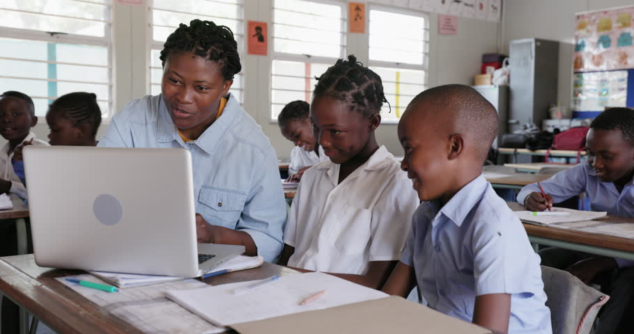 Close-up. Black African female teacher sitting and assisting African children at their desk with a task on a laptop in a classroom in Africa Royalty-Free Stock Footage #1108928325