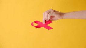 Vertical video of female hand holding small red ribbon, posing isolated on pastel yellow studio background wall with copy space for promotion content or design. HIV awareness concept, world AIDS day - Powered by Shutterstock - Get 15% off with code: PIKWIZARD15
