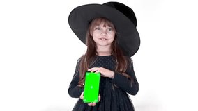 Cute little smiling witch in a black hat holding a greenscreen phone in her hands. Halloween Footages. Chroma key smartphone set up for advertising. High quality FullHD footage