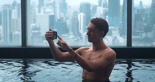 Man 30s call video online on mobile phone at hotel swimming pool on vacation. Shirtless male have video chat at indoor pool of skyscraper. Smiling tourist man having video call in luxury hotel pool