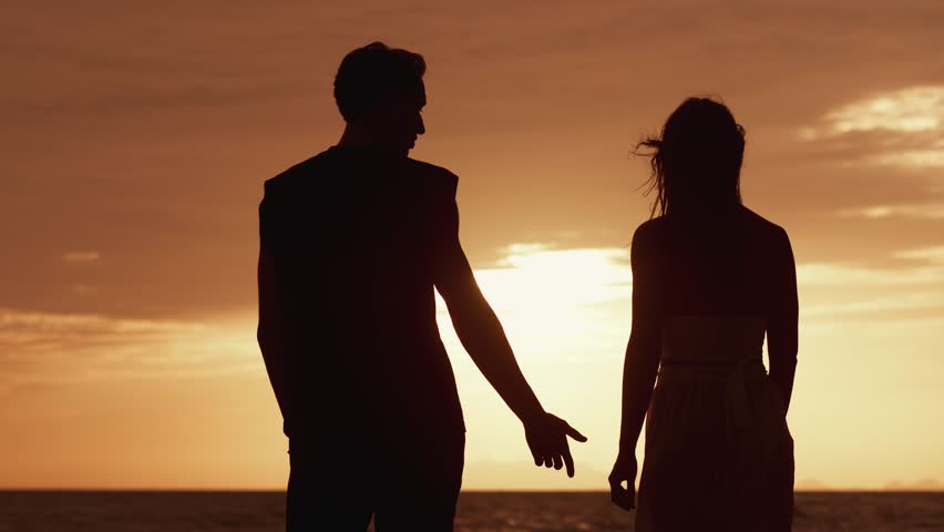 Rear view of man in love takes a woman's hand on a date against an orange sunset. Silhouette of a loving couple at sunset on the sea during a date. Concept of love