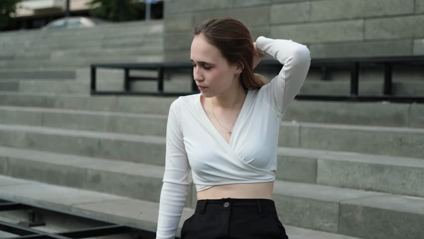 A cute teenage girl with long brown hair, wearing a white top, sits on the steps on a warm summer day.